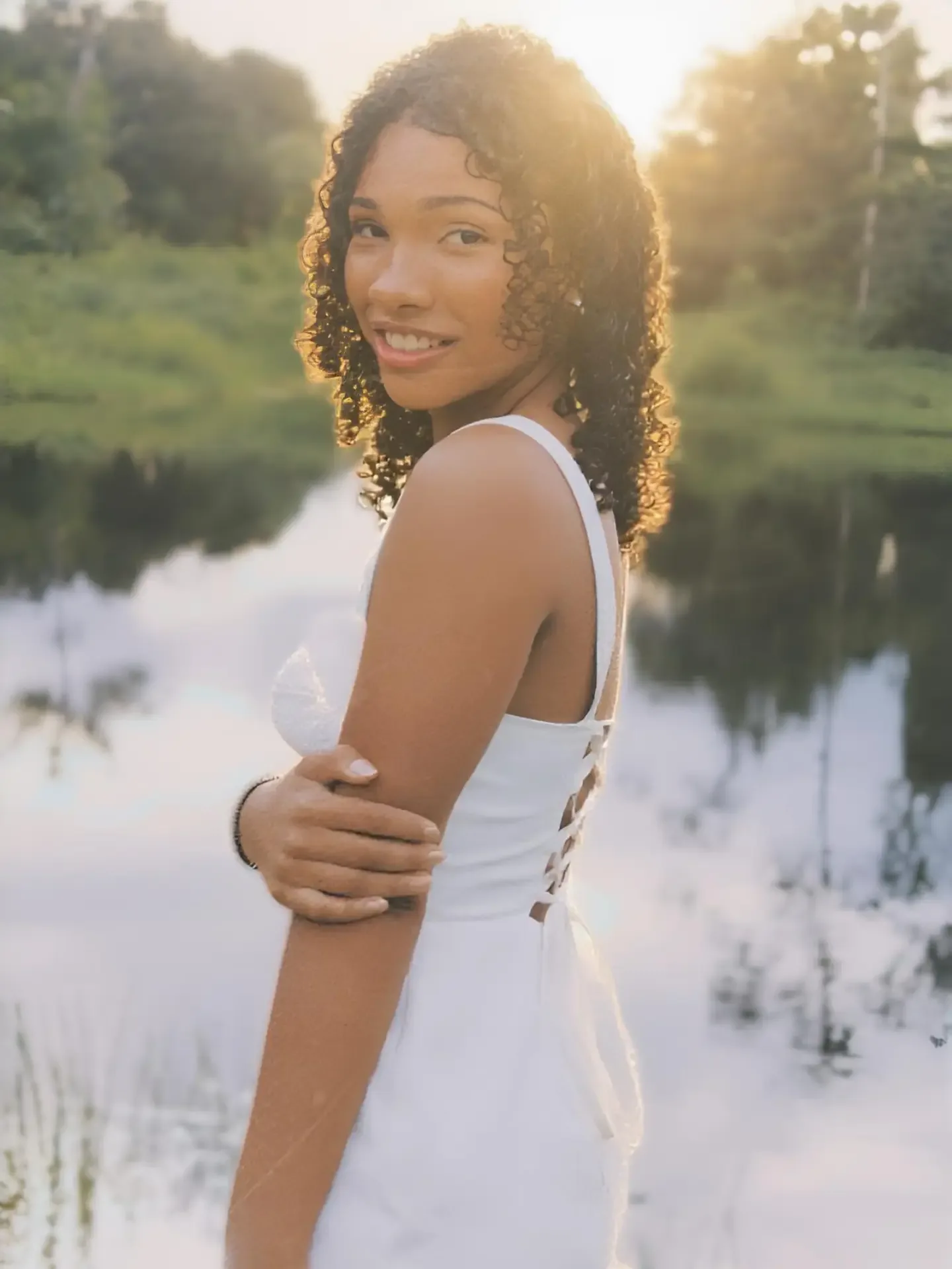 Mulher de vestido branco olha por cima do ombro à beira de um lago, com o sol ao fundo.