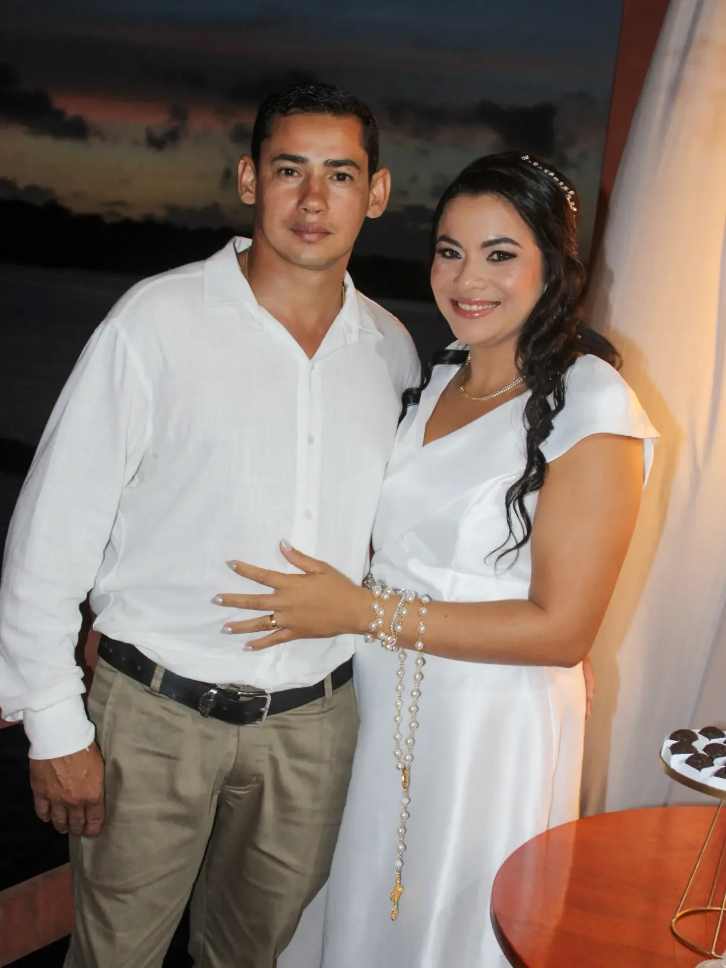 Noivos vestidos de branco posam abraçados; ele apoia a mão na cintura dela.