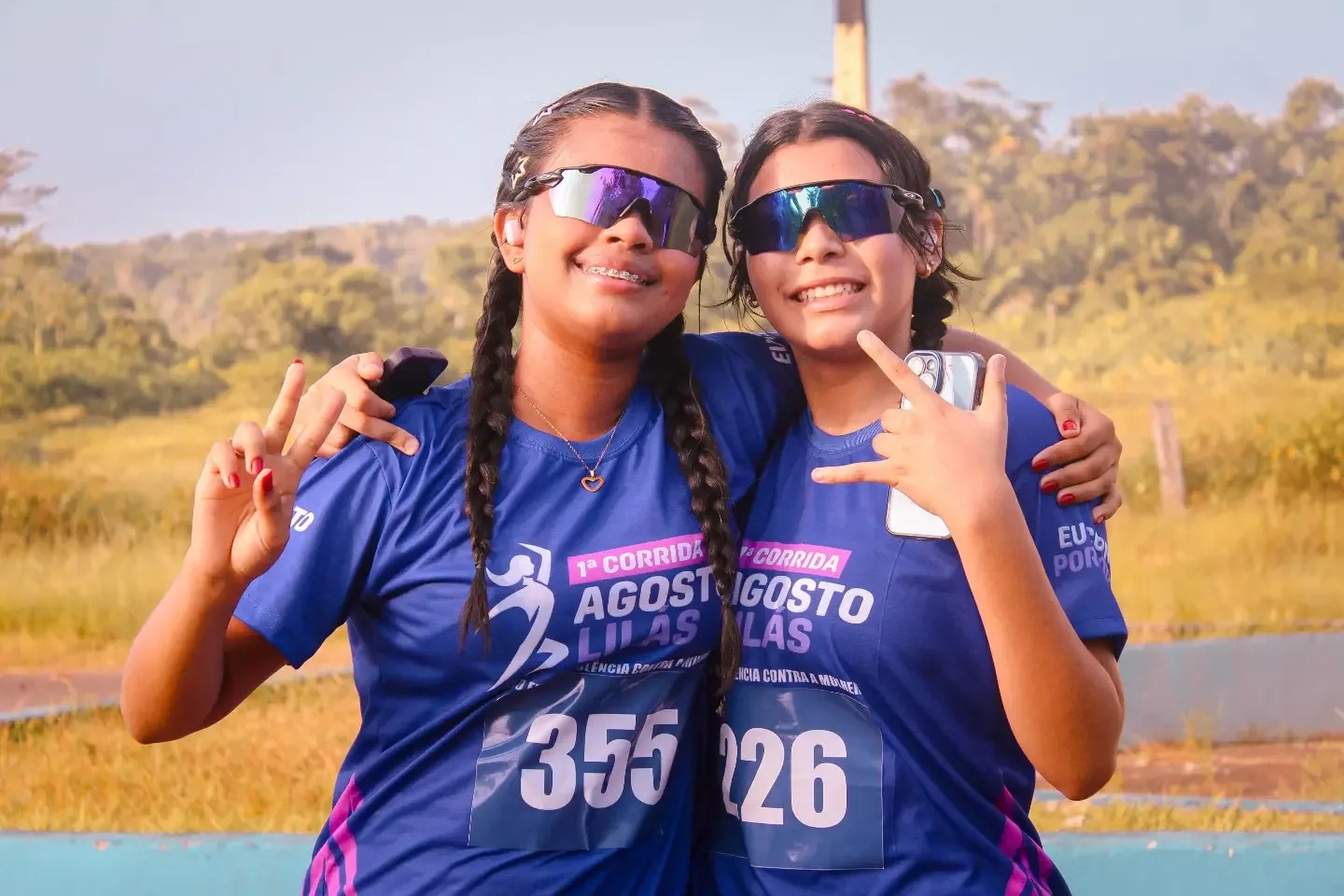 Duas corredoras abraçadas, de óculos escuros, com camisetas e números de prova.