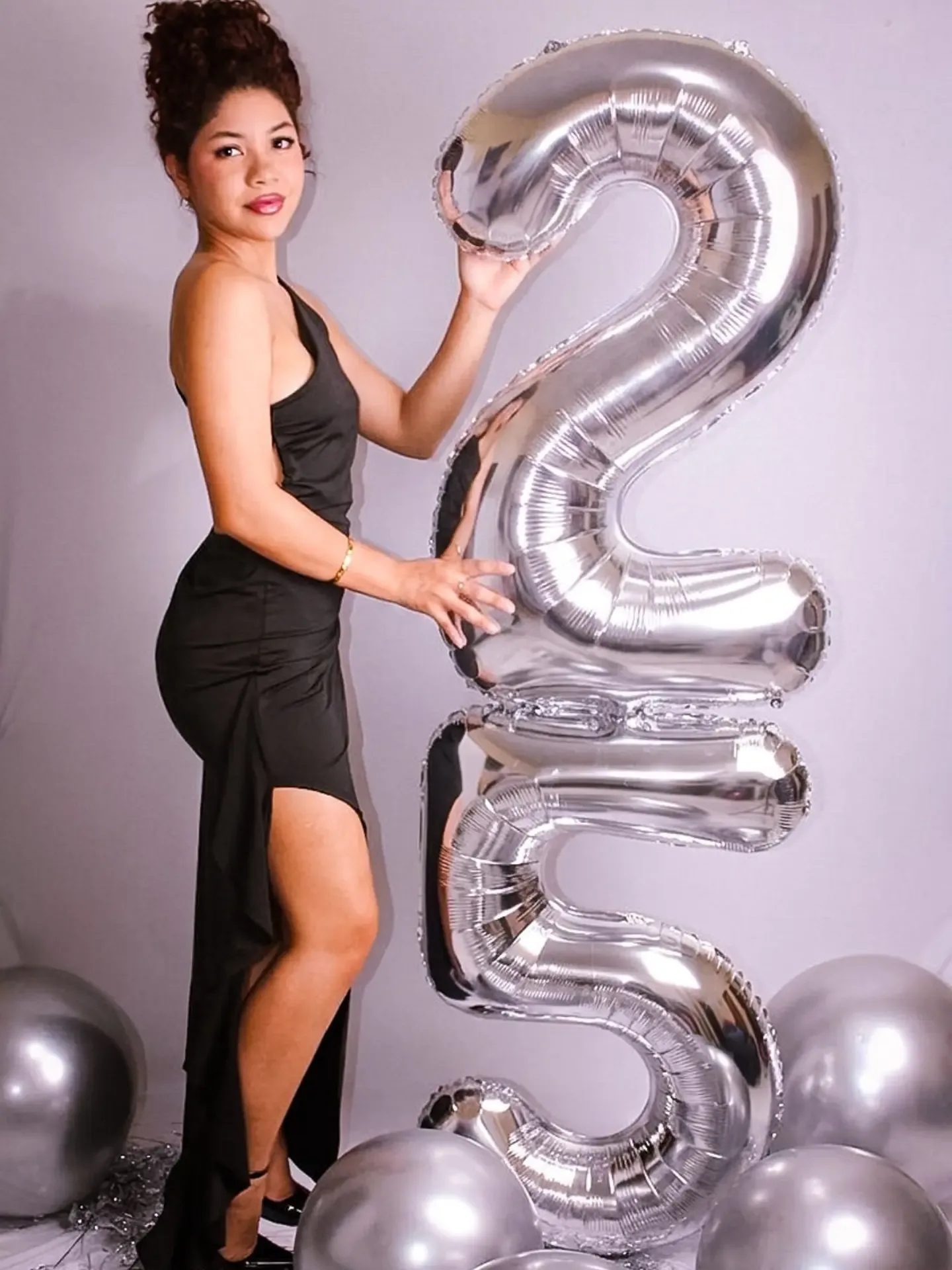 Mulher de vestido preto apoiada em balões prateados com os números 2 e 5.