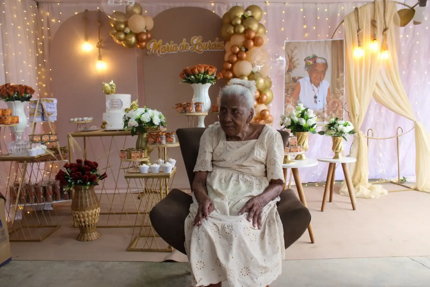 Senhora de vestido bege sentada em uma poltrona, em festa decorada com balões dourados.