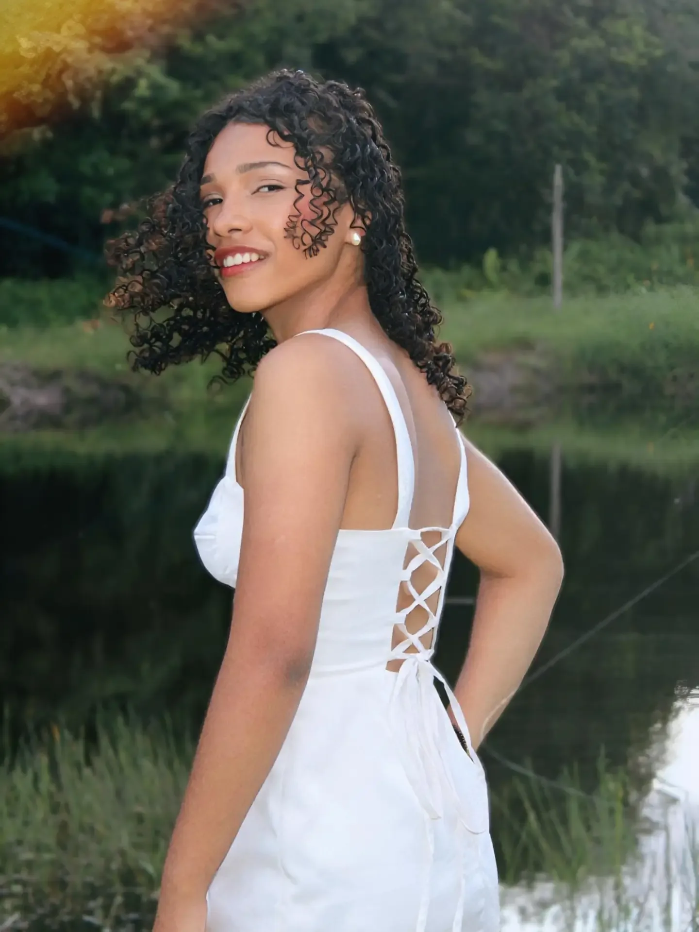 Mulher de vestido branco, de costas, olha por cima do ombro à beira de um lago.
