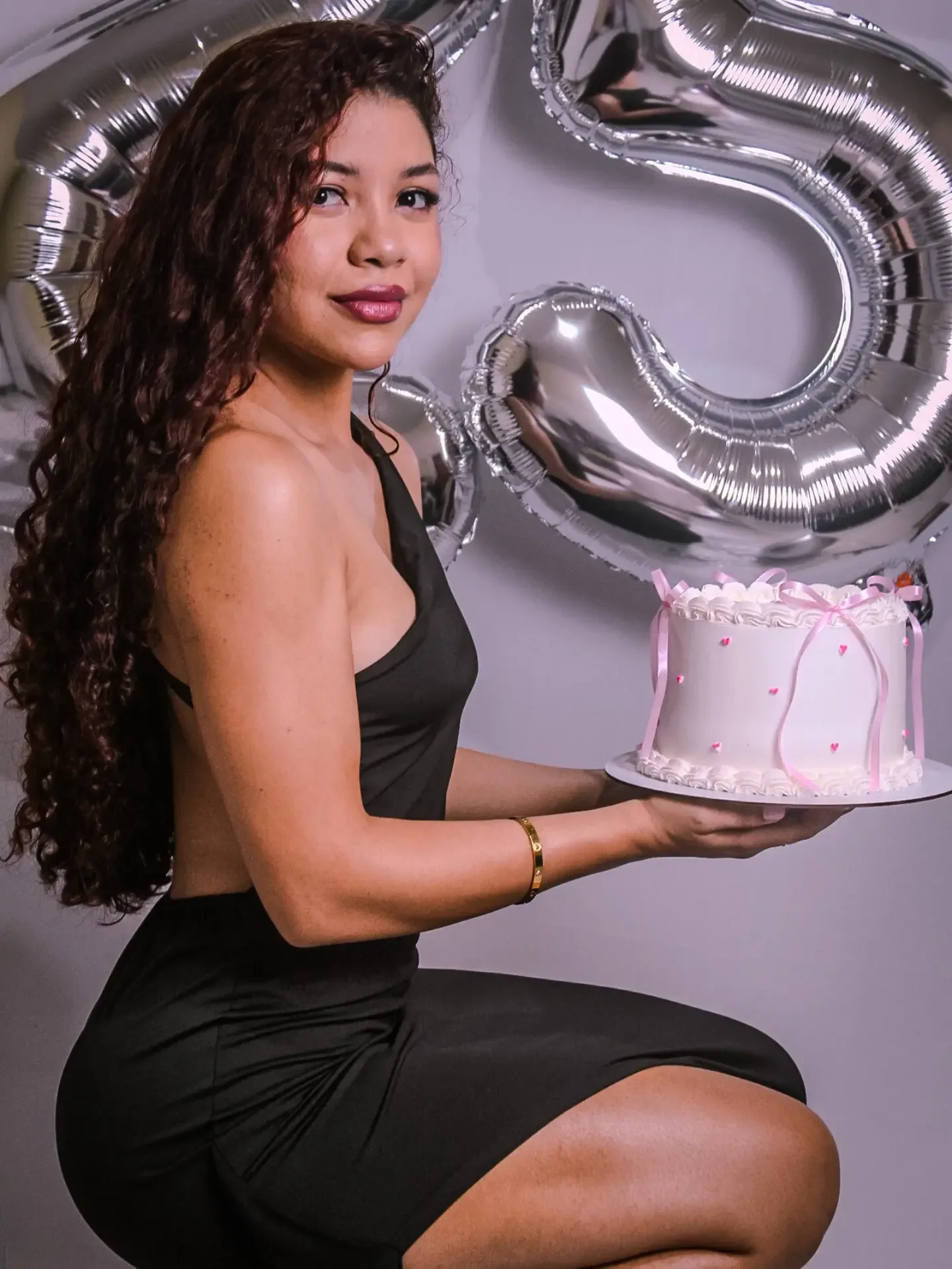 Mulher de vestido preto ajoelhada segura um bolo branco, com balões prateados do número 25 ao fundo.