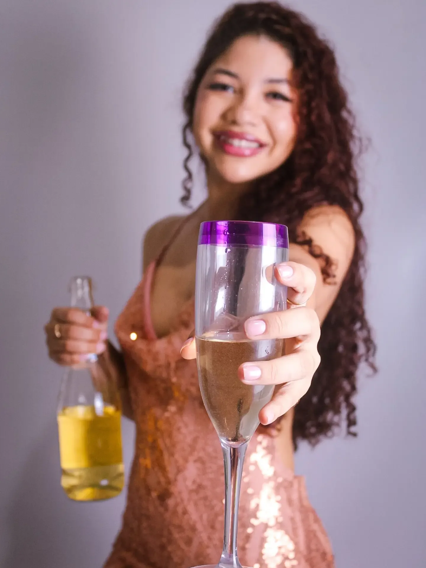 Mulher de vestido rosé brilhante estende uma taça de espumante para a câmera.