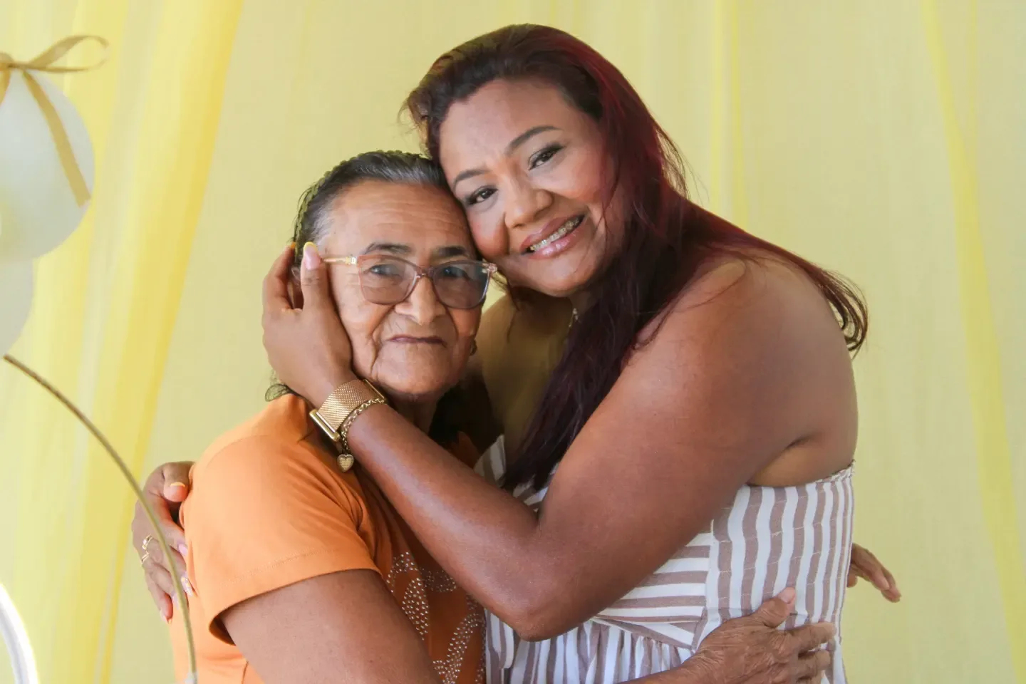 Duas mulheres abraçadas, rosto com rosto, em uma festa de aniversário.