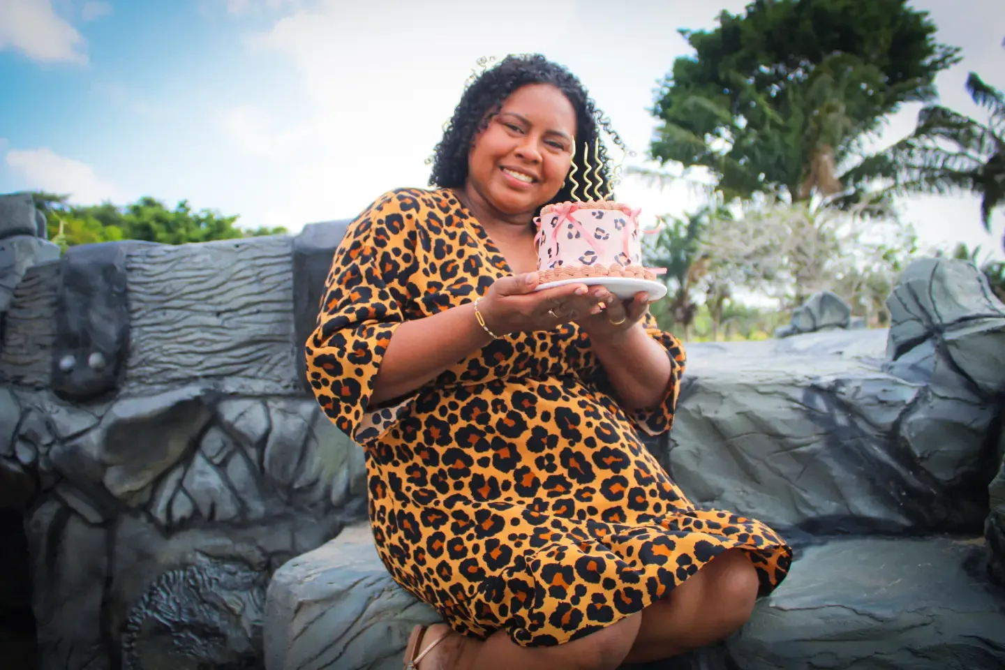 Mulher de vestido de oncinha segura um bolo, sentada sobre pedras ao ar livre.