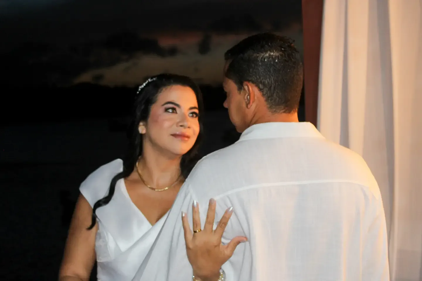 Noiva mostra a aliança com a mão no ombro do noivo, ao entardecer.