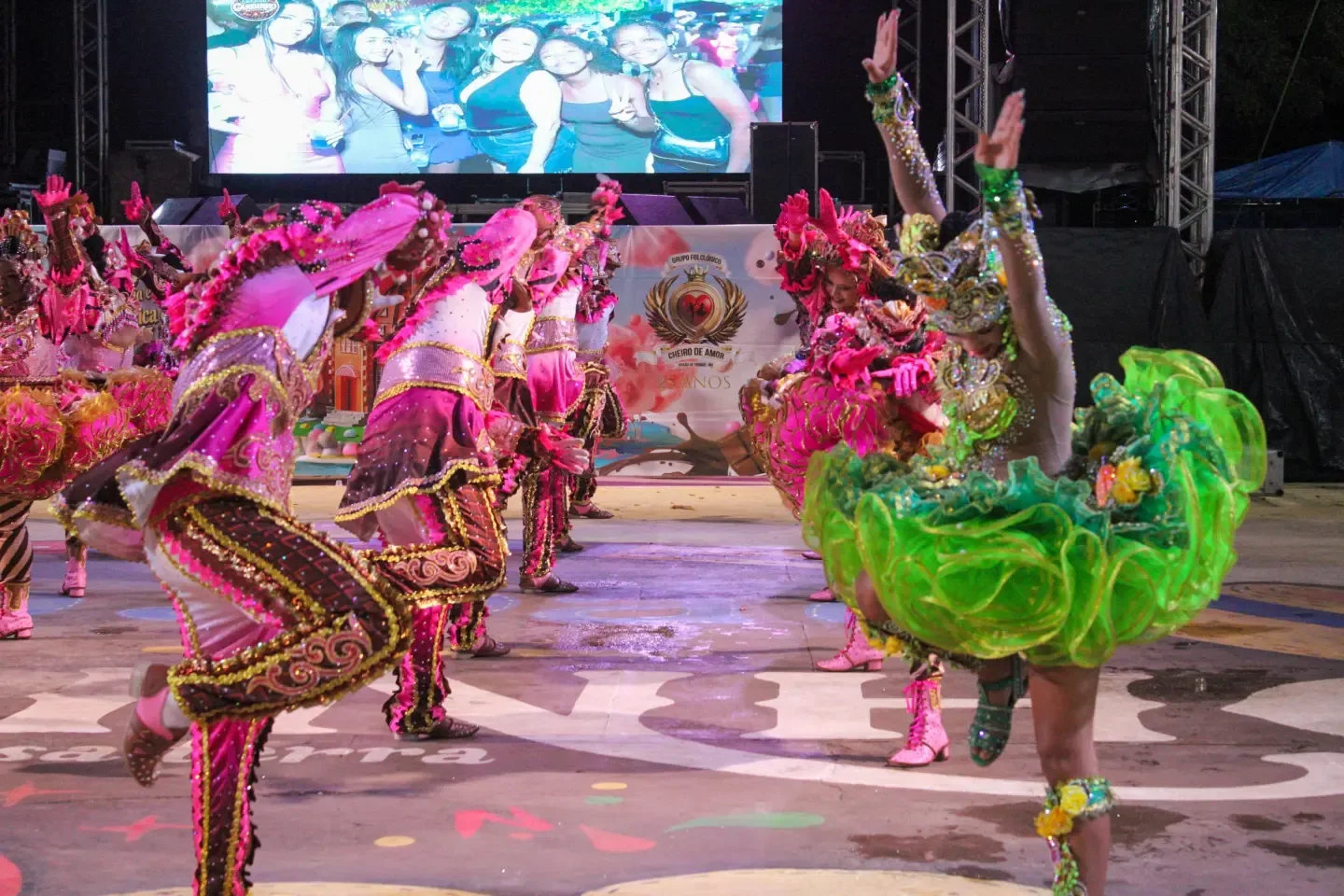 Grupo de dançarinos com figurinos rosa se apresenta diante de um palco iluminado.