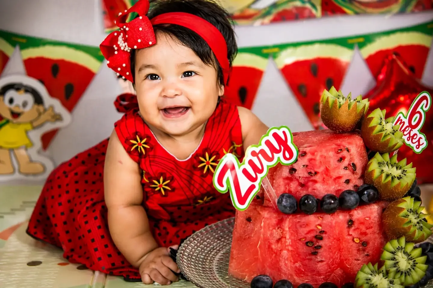 Bebê de vestido vermelho sorri ao lado de um bolo de melancia decorado com frutas e uma placa de 6 meses.