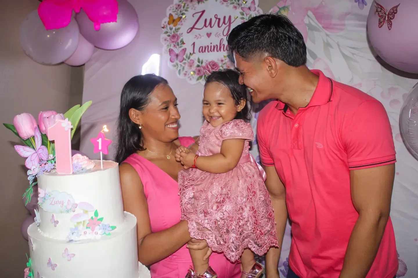 Bebê de vestido rosa no colo dos pais diante do bolo de aniversário de 1 ano.