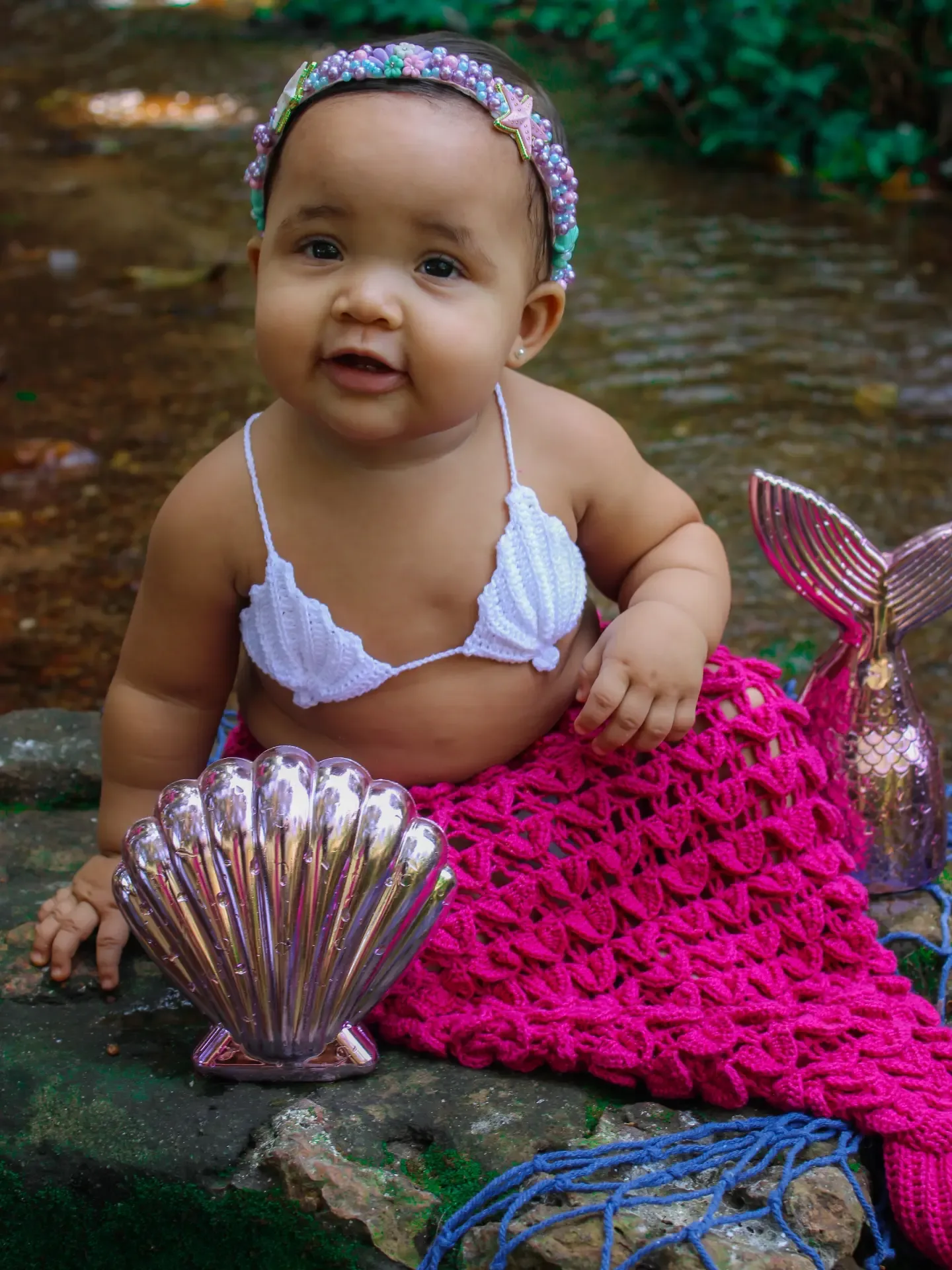 Bebê com cauda de sereia rosa de crochê sentado sobre pedras.
