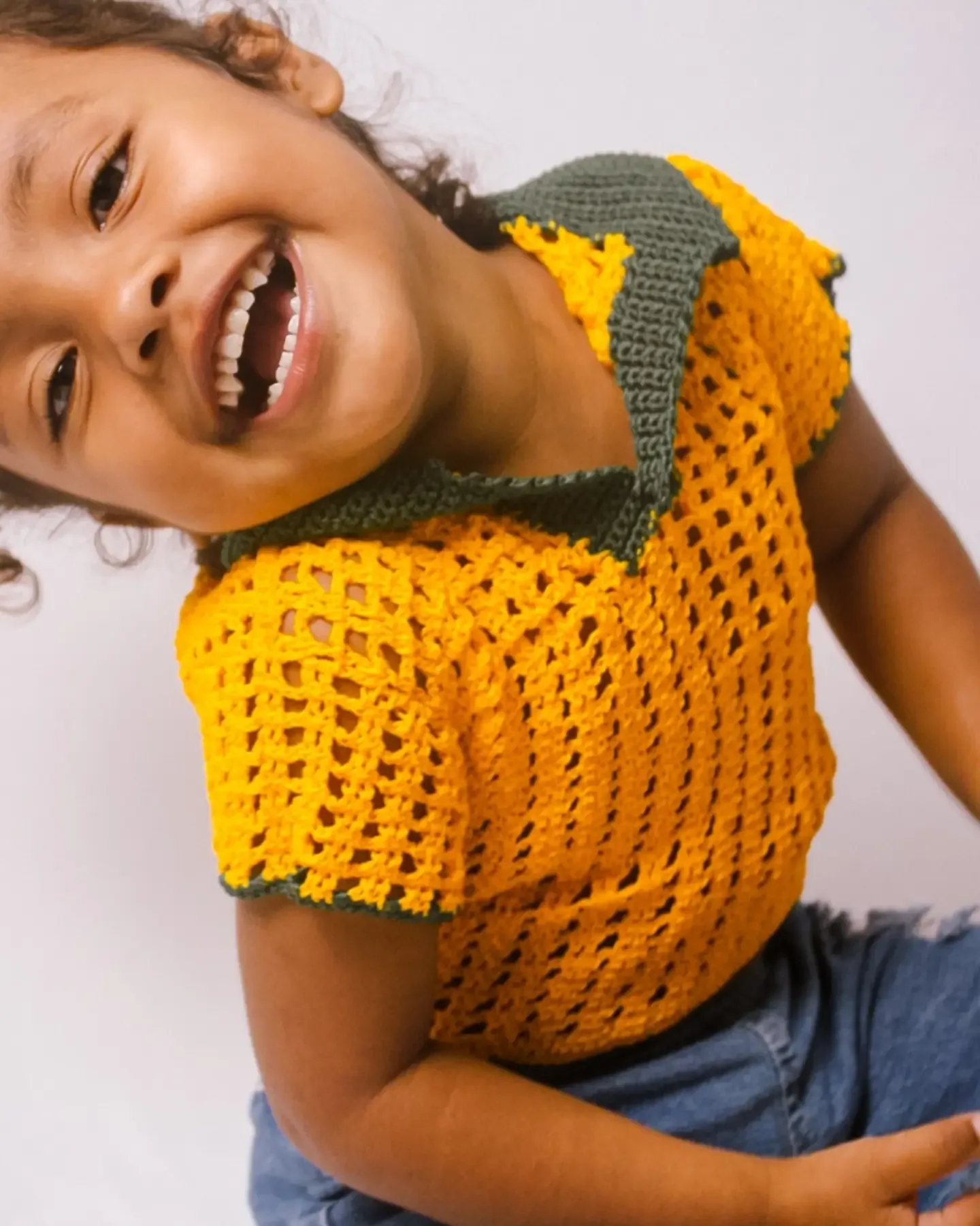 Criança de blusa de crochê amarela ri, inclinada para a câmera.