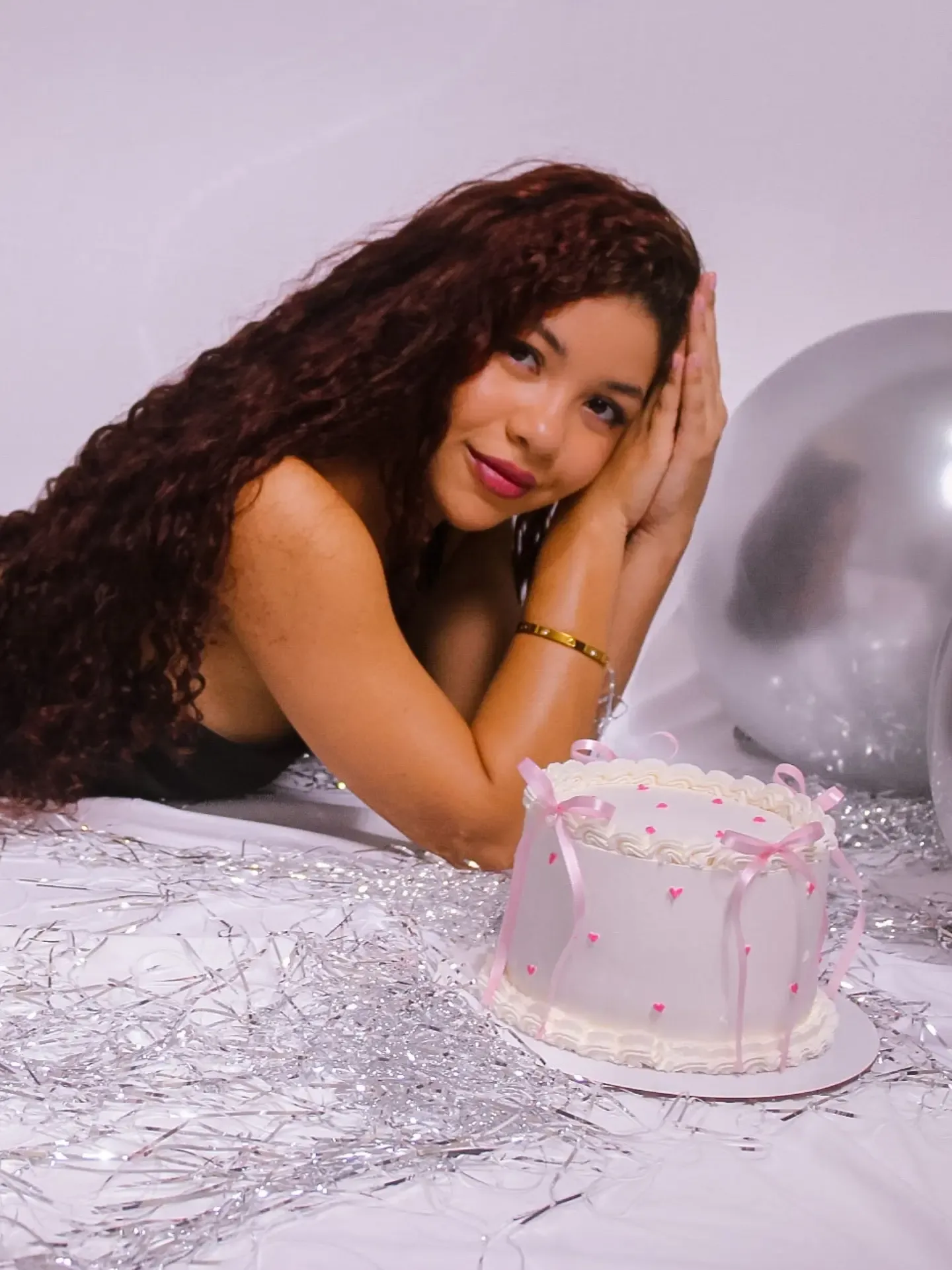 Mulher de cabelo longo e cacheado deitada ao lado de um bolo branco, com as mãos no cabelo.