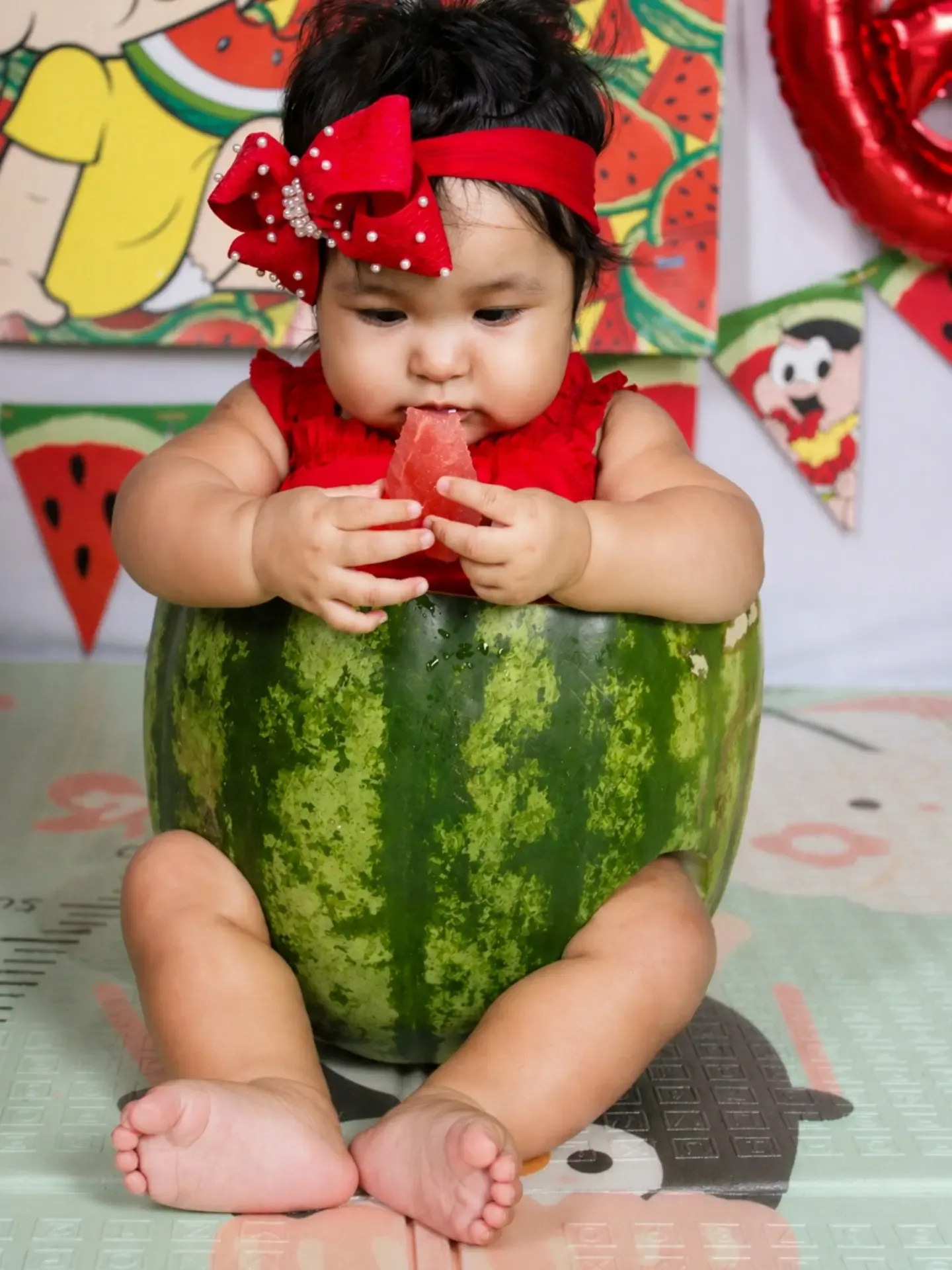 Bebê sentado dentro de uma melancia cortada, em cenário vermelho.