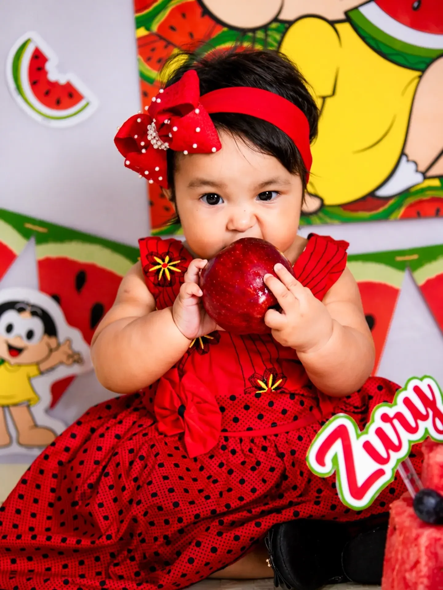 Bebê de vestido vermelho e laço na cabeça morde uma maçã em cenário de melancias.